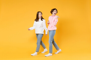 Fototapeta na wymiar Full length side view of excited shocked two young women friends 20s wearing casual white pink hoodies walking going pointing thumb aside isolated on bright yellow color background studio portrait.