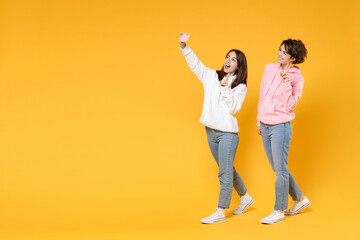 Full length of excited two young women friends 20s wearing casual white pink hoodies doing selfie shot on mobile phone showing victory sign isolated on bright yellow color background studio portrait.