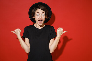 Shocked amazed young brunette woman 20s years old wearing casual basic black t-shirt hat standing pointing thumbs aside on mock up copy space isolated on bright red color background studio portrait.