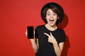 Shocked young brunette woman 20s in casual basic black t-shirt hat standing pointing index finger...