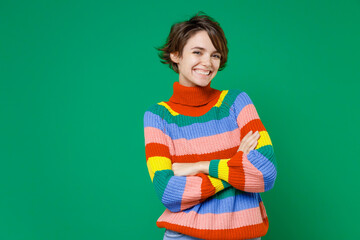 Smiling beautiful young brunette woman 20s years old wearing basic casual colorful sweater standing holding hands crossed looking camera isolated on bright green color background studio portrait.