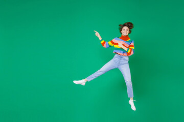 Fototapeta na wymiar Full length of smiling cheerful funny young brunette woman 20s in colorful sweater jumping pointing index fingers aside on mock up copy space isolated on bright green color background studio portrait.
