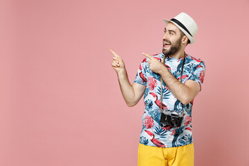 Excited young traveler tourist man in summer clothes hat with photo camera point index fingers aside up isolated on pink background. Passenger traveling abroad on weekends. Air flight journey concept.