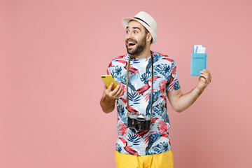 Excited young traveler tourist man in summer clothes hat using mobile cell phone hold passport tickets isolated on pink background. Passenger traveling abroad on weekends. Air flight journey concept.