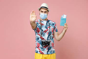 Dissatisfied traveler tourist man in mask to safe from coronavirus hold passport tickets showing stop gesture isolated on pink background. Passenger traveling on weekend. Air flight journey concept.