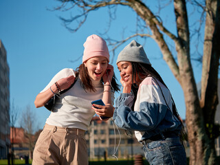 two beautiful girl friends sharing a smart phone through earphones, wearing casual clothes and looking happy