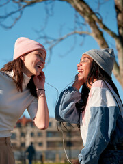 two girls looking happy while sharing a smartphone and earphones, outside and wearing casual clothes