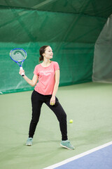 A young white brunette girl with a racket in her hands and in black sweatpants and a pink T-shirt is playing tennis and smiling. It is located in the indoor table tennis hall.