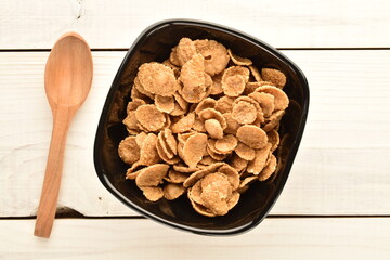 Natural multi-grain flakes in a black ceramic plate with a wooden spoon on a white wooden table, close-up, top view.
