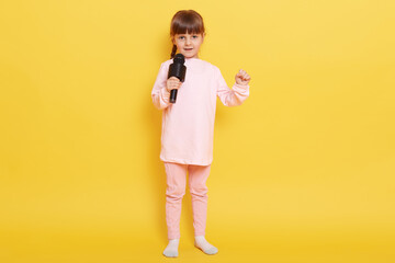 Close up photo of cool attractive lady kid having fun, standing isolated over yellow background, adorable kid with microphone arranging concert, dresses pale pink background.