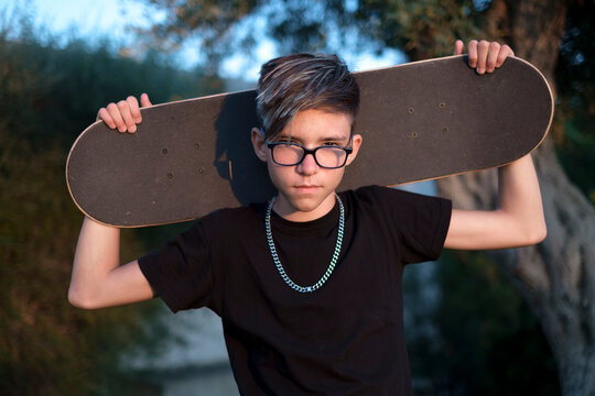 Adolescente con gli occhiali guarda serio tenendo dietro la testa il suo skateboard