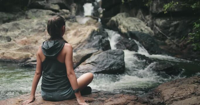 Thailand's river waterfall: woman rest on stones. Calm water flow on stream rock bottom. Girl relaxing back view: National Park Than Sadet, Koh Phangan Island. Cinematic footage shot in 4K, UHD