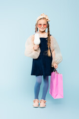 full length of little girl in sunglasses holding glass with milkshake and shopping bag on blue