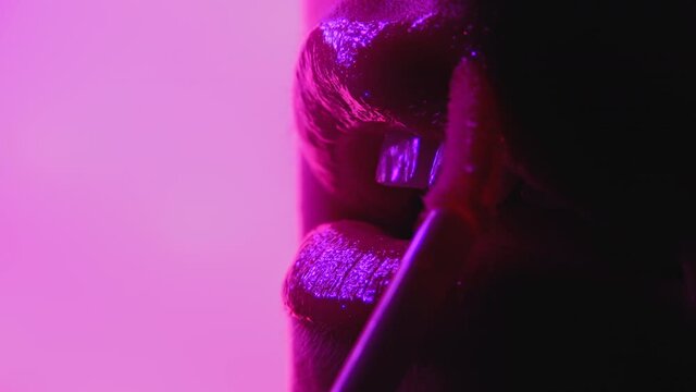 Woman applying liquid gloss or lipstick on her sexy lips. Flickering colorful neon light. Extreme close up sensual female face. Advertising make-up, beauty, cosmetics concept