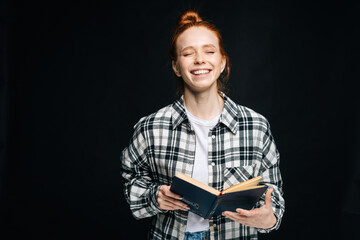 Naklejka premium Laughing young woman with closed eyes college student holding opened book on black isolated background. Pretty redhead lady model emotionally showing facial expressions in studio, copyspace.