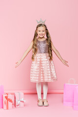 full length of happy little girl in crown standing near presents and shopping bags on pink