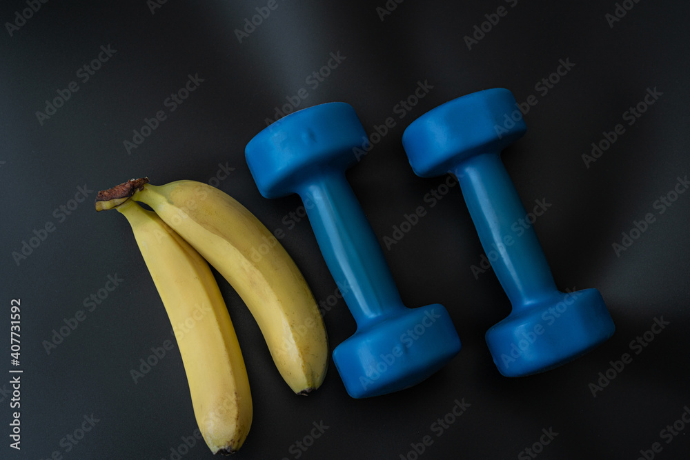 Wall mural two blue dumbbells for fitness lie on a black background, and in front of them are two bananas.