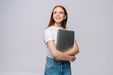 Smiling young woman student holding laptop computer and looking away on isolated gray background. Pretty lady model with red hair emotionally showing facial expressions in studio, copy space. - obrazy, fototapety, plakaty