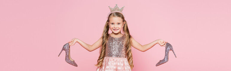 cheerful little girl in crown and dress holding heels isolated on pink, banner