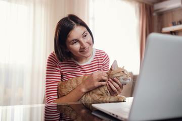 Happy young woman works at home with a laptop and a ginger cat