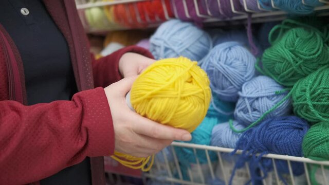 Women hands in the store choose bright yellow yarn for knitting and needlework. In the background you can see shelves with colorful wool