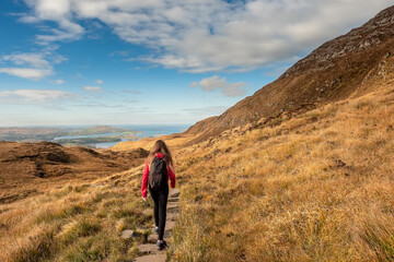 Fototapeta premium Teenager girl walking on a path by a mountain. Beautiful scenery in the background. Connemara National park, county Galway, Ireland. Warm sunny day, Cloudy sky. Outdoor activity and tourism concept