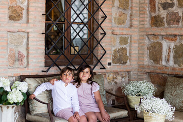 niña y niño vestidos elegantes para celebración, sentados en bonito banco, bajo ventana con reja...