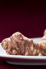 Detail of single Jerusalem artichokes (Helianthus tuberosus), also known as sunroot, sunchoke, or earth apple, lay on a white plate against a scarlet background. Vertical frontal view with text space