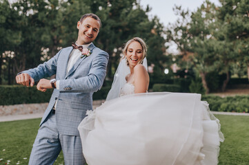Stylish groom in a gray checkered suit and a blonde bride in a long white dress are cheerfully and...