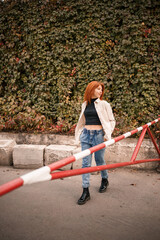 Fototapeta premium A pretty redhead young woman is walking down the street, she is dressed in jeans and a beige shirt. Beautiful girl dressed in casual style with a smile on her face
