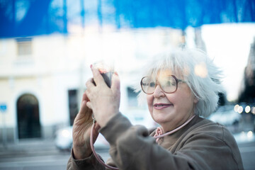 elderly woman taking a selfie in the street behind glass with reflections from car lights