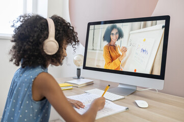 Homeschooling and technology concept. Cute clever schoolgirl fills exercise book in watching online video classes, webinars, tutorial with a African female teacher on the PC screen.
