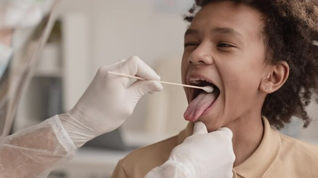 Close-up of teenage African boy opening his mouth for unrecognizable medical worker collecting sample of his saliva with cotton swab for analysis. Patient smiling and nodding