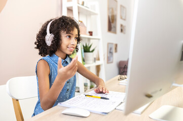 Cute curly African schoolgirl in headphones is talking online with classmates, smiling little girl participates in shared lesson on the PC studying from home. Homeschooling, e-study concept