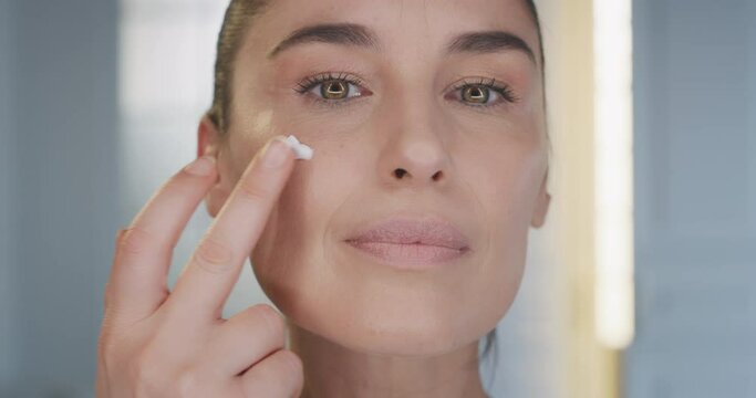 Beauty shot of mature woman applying moisturizing anti wrinkle day or night cream on her previously cleaned face and smiling in camera on blue background. Concept of skincare, cosmetics, healthcare.