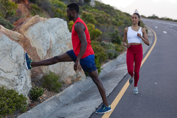 Fit african american man in sportswear stretching while woman is running on a coastal road