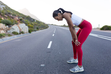 Fototapeta na wymiar Fit african american woman in sportswear standing and resting on a coastal road
