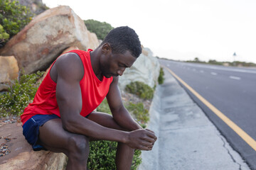 Fit african american man in sportswear sitting on rock by a coastal road