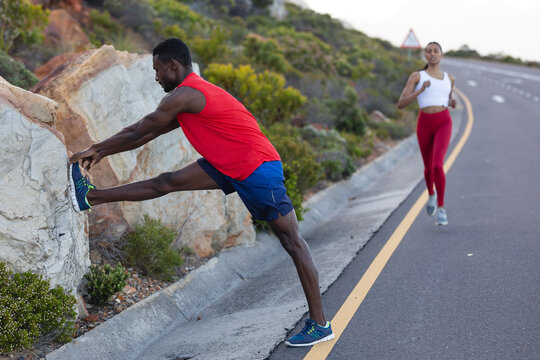 Fit african american man in sportswear stretching while woman is running on a coastal road - Powered by Adobe
