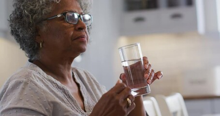 Senior african american woman taking medication and drinking water while sitting at home - Powered by Adobe