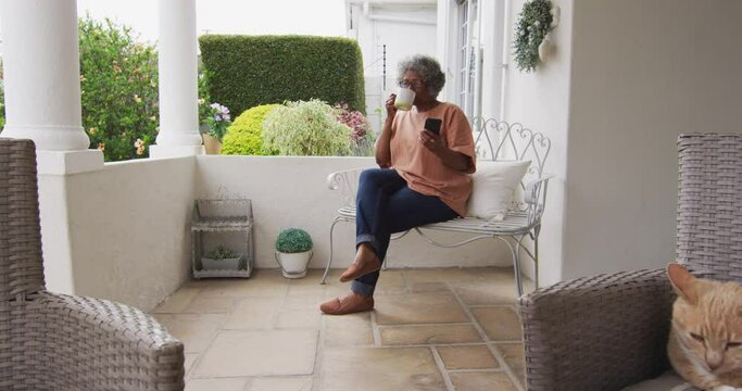 Senior african american woman using smartphone while drinking coffee sitting on the porch of the hou