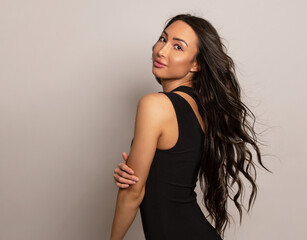 Beautiful young woman with long shiny black hair and natural make-up posing in dress on grey background 