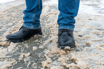 A man stands on the sidewalk in snow and mud. Black shoes and blue jeans close-up on the background...