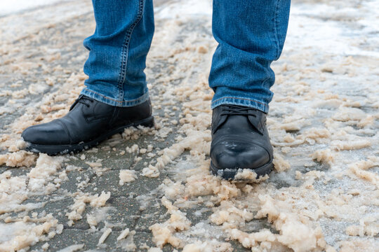 A man stands on the sidewalk in snow and mud. Black shoes and blue jeans close-up on the background of dirty snow. Ice on the road and sidewalk, the use of reagents, sand and salt.