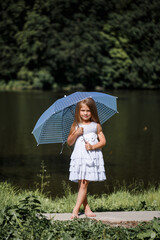 A little girl with long hair in a white dress stands on the bank of the river. Walk by the lake on a summer sunny day