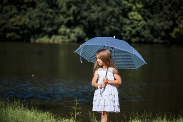 A little girl with long hair in a white dress stands on the bank of the river. Walk by the lake on a summer sunny day