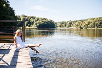 A 7-year-old girl with long blond hair by the lake sits on a clutch with legs in the water. She splashes her feet in the lake. Barefoot girl in a white dress with long hair.