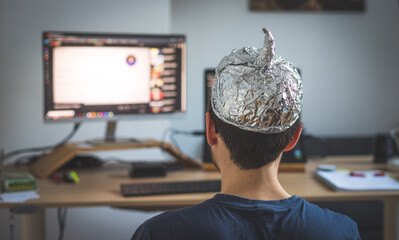 Conspiracy theory concept: young man is wearing aluminum head, sitting in front of the pc watching...