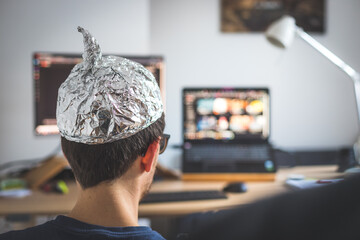 Conspiracy theory concept: young man is wearing aluminum head, sitting in front of the pc watching videos - obrazy, fototapety, plakaty