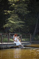 Family, mother and daughter are sitting by the river. Little girl with her mom. Girls in white dresses. Family time together.